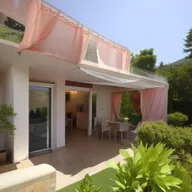 A two-story house on a 240m² property. Ground floor features two bedrooms, a living room, dining room, and an American kitchen. Living room curtains part to reveal a garden with a water fountain. First floor enjoys two bedrooms and a balcony overlooking the living room