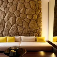 A spacious 4m by 8m room featuring a massive natural rock wall with heavy relief, subtly lit by indirect light and a mirror seamlessly incorporated. Dominating the room is a large, minimalist white sofa adorned with yellow pillows, complemented by a Karim Rashid coffee table.