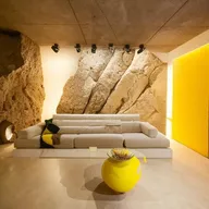 A spacious 4m by 8m room featuring a massive natural rock wall with heavy relief, subtly lit by indirect light and a mirror seamlessly incorporated. Dominating the room is a large, minimalist white sofa adorned with yellow pillows, complemented by a Karim Rashid coffee table.