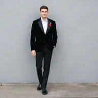 A man dressed for a school farewell, wearing a black velvet blazer over a black shirt, black pants, and matching black formal shoes. His look is embellished with an artificial red flower in his pocket and a sleek black watch.