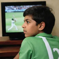 A boy wearing a shirt with the name 'Zaheer Abbas' is engrossed in watching a T20 cricket match between Pakistan and New Zealand on a sleek, modern LED television.